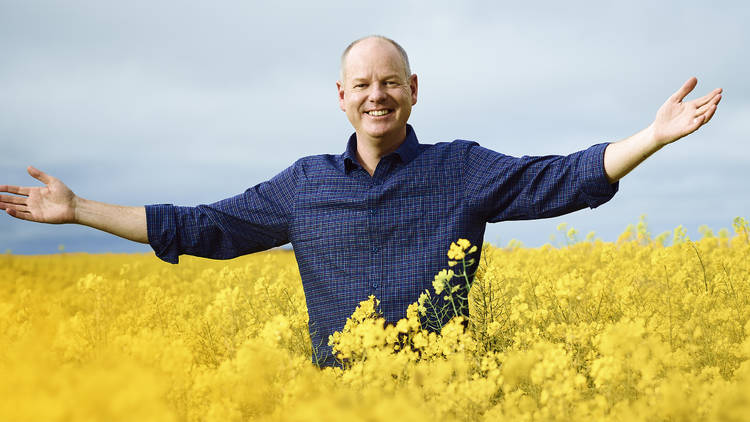 Tom Gleeson, wearing a blue checkered shirt, standing arms outstretched in a fields of bright yellow flowers.