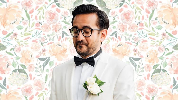 Comedian Sami Shah in white wedding tux in front of pastel floral wallpaper