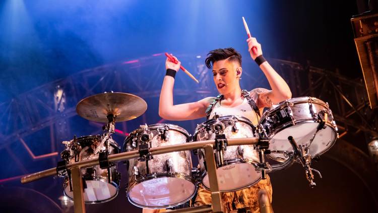 A woman wearing a white singlet, black wrist cuffs and a short spiky hair cut looms over a drum kit, mid performance