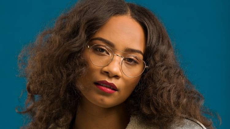 A woman with thick, dark wavy hair, red lipstick and wire rimmed glasses looks straight ahead