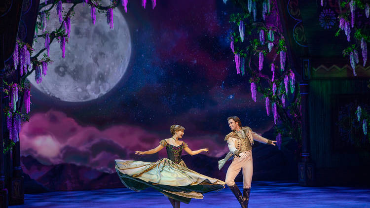 A woman in a ballgown twirls with a man wearing a suit on a stage. The set features a large moon and hanging wisteria flowers