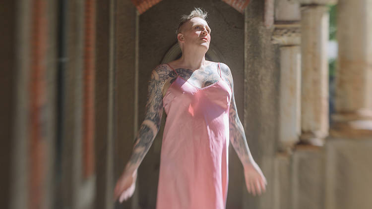 A woman with short blonde hair and tattoos across their chest and arms wears a pale pink slip dress and looks serenely up while light touches them