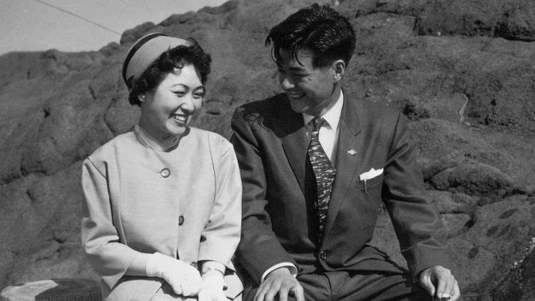 In a black and white photo a couple sits in front of a landscape wearing vintage attire, the woman smiles coyly and the man looks at her adoringly.