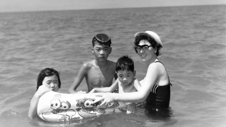 In a black and white photo a woman and three children are at the beach, they are in the water with a floatie device