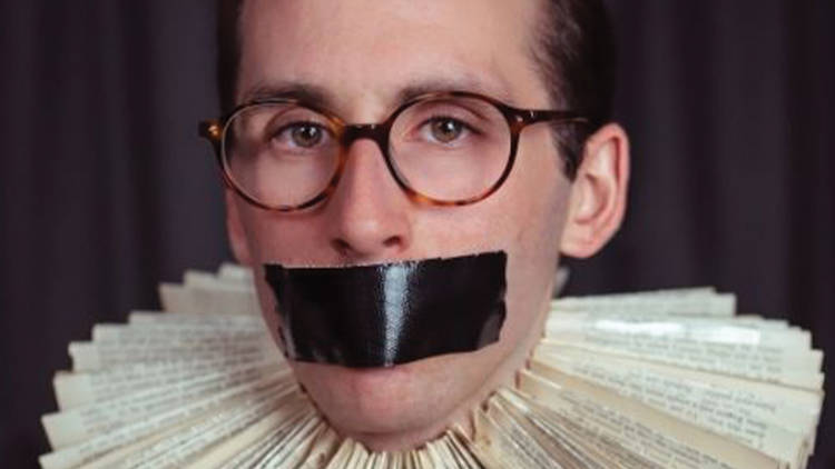 A man wearing tortoiseshell circular glasses stares straight ahead with black gaffer tape over his mouth. He is wearing a neck ruff made of book pages