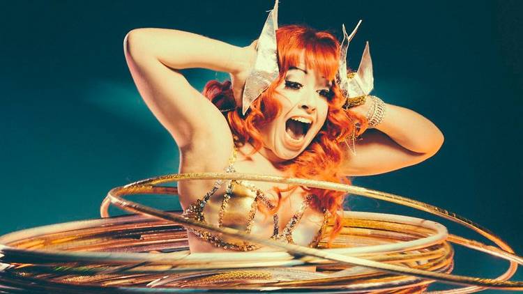 Performer with curly orange hair and gold costume is hula hooping with a stack of gold hoops around their waist, they have their hands pressed to the sides of their head with a wild expression.