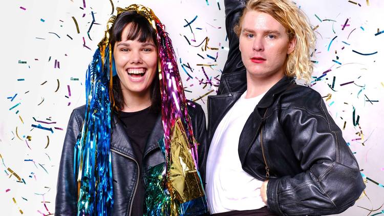 Two people surrounded by falling rainbow coloured shiny confetti. Both are wearing black leather jackets. 