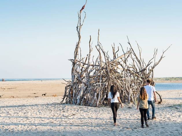 Gippsland road trip guide - Visit Victoria People admire a giant pile of driftwood fashioned into a pointed hut or structure on a white sand beach.