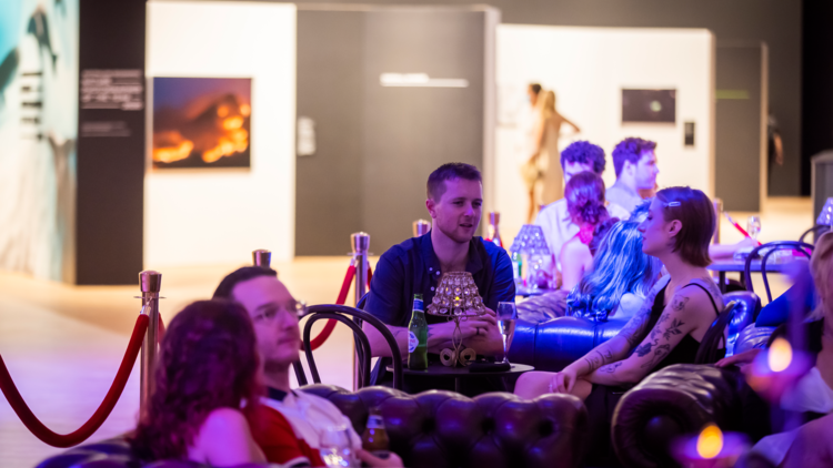 Lots of people sitting on leather couches in purple lighting. A photographic exhibition can be seen in the distant background.