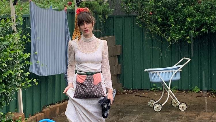 Comedian Beth McMullen poses in front of clothesline in white lace dress, wears pouch around waist.