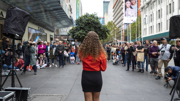 A woman with a red jumper, black skirt and long curly brown hair performs to a crowd in Bourke Street Mall.