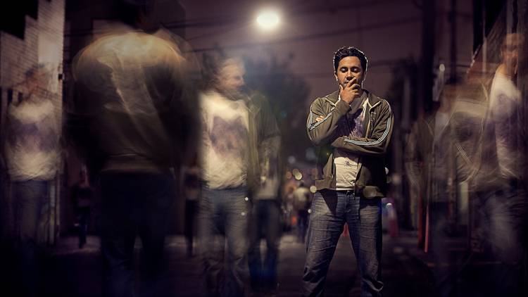 A man stands thinking under a streetlamp at night. Ghosts of other people are passing by.