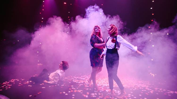 Two women dance in pink smoke while one lies on the floor in petals