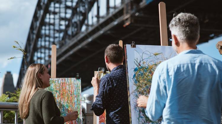 People with easels create scribbly drawings, they hold wine glasses, the Sydney Harbour Bridge is in the background.