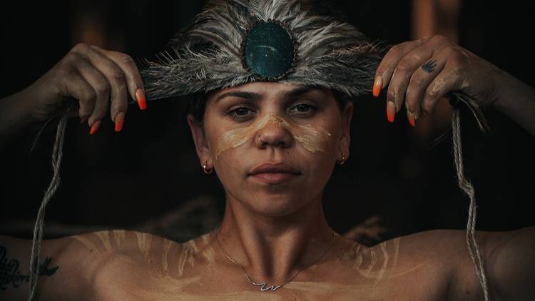 A woman with bright red nails and markings painted on her face and shoulders puts a feathered headband up to her forehead