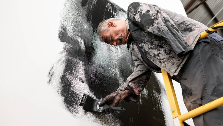 An elderly man with short white hair and only one warm meditatively smears black paint on a white wall. He is covered in black paint too.