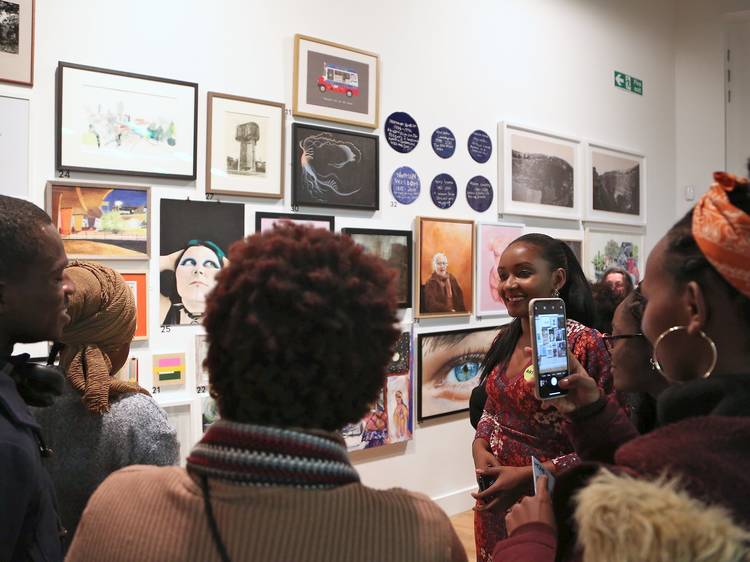 A group of people photograph an artist on their phones as she stands in front of a wall of artworks