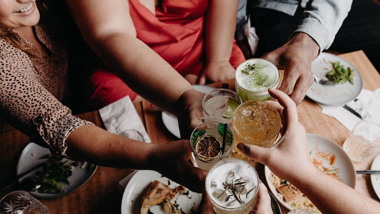 Five people cheers in the centre of the photo with five different drinks.