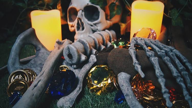 A skeleton lies between two large candles. Beneath it are jewels and coins.