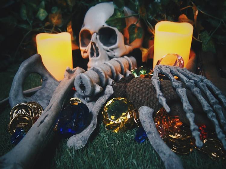 A skeleton lies between two large candles. Beneath it are jewels and coins.