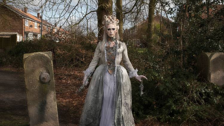 A ghostly figure in pale make up and gown stands in a wood next to a large stone gatepost 