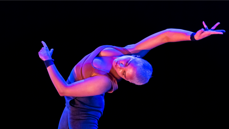 An outstretched woman with cropped blonde hair is bathed in purple and pink light 