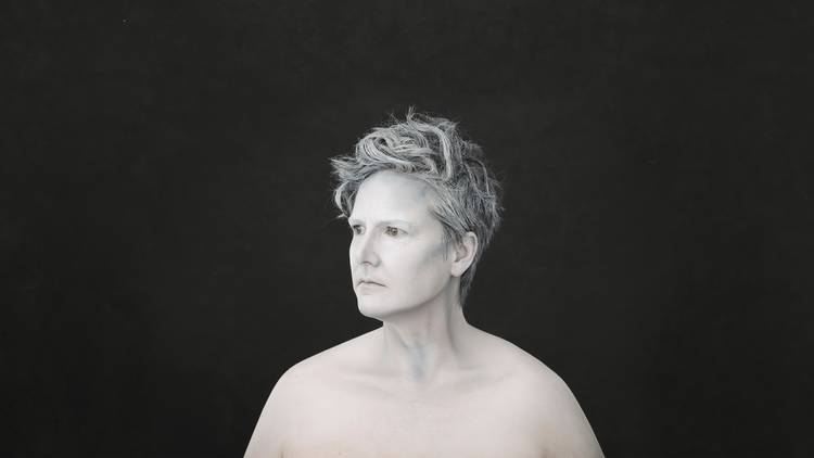 A black and white portrait photograph of Hannah Gadsby taken from the shoulders up.. She is stoically turning her face to the side and is wearing wearing no shirt. She appears like a marble statue.