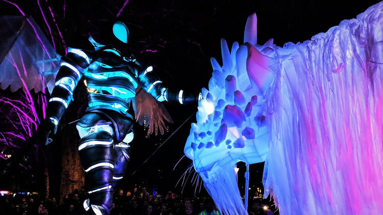 A giant glowing astronaut-like puppet stands next to a ten-metre-long crystalline lion puppet, also glowing. It is nightime.