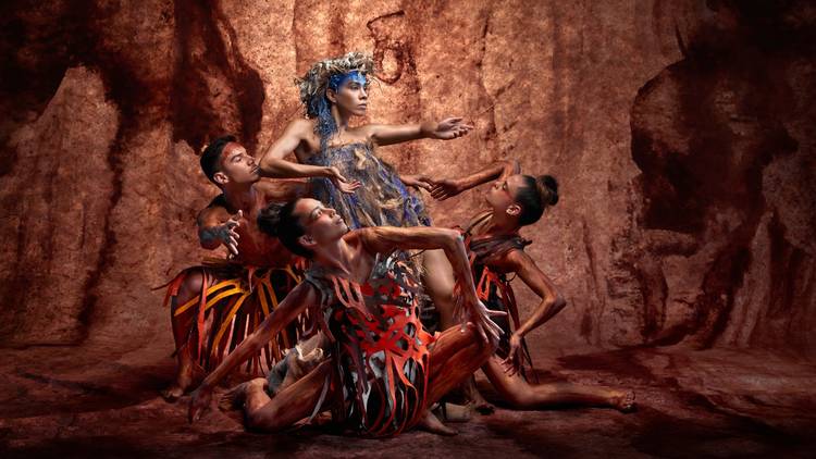 A group of people in earthen colours create a pyramid of bodies against a red rocky backdrop