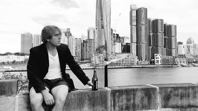 A man with no pants cradles a beer in front of Sydney city skyline