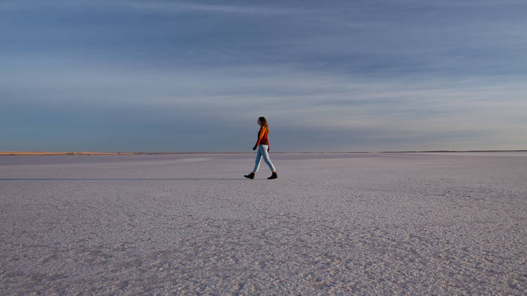 A woman wearing jeans and an orange sweater walks across a salt lake under a vast blue sky