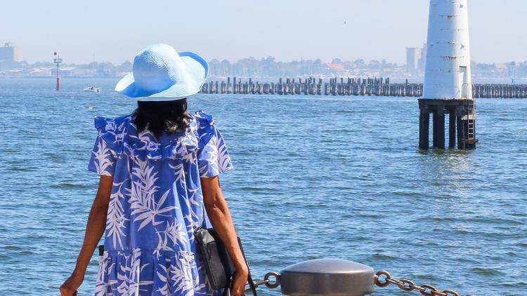 A woman faces the ocea while wearing a sun hat and blue dress. She is carrying a lot of luggage