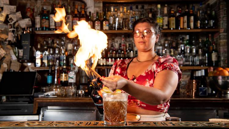 A person holds a large flame over a cocktail at a bar.