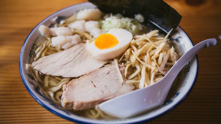 A bowl of ramen in a blue and white china bowl. There is a white spoon in the bowl and the ramen is topped with two thick slices of pork, half a soft boiled egg and a slice of dried seaweed