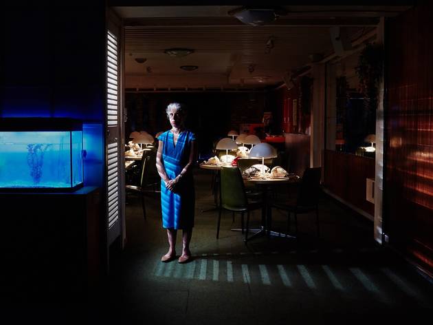 The Nightline / Rising A woman in a blue dress stands half lit in a dark room. The room is filled with tables that have lamps on them