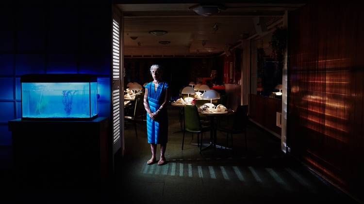 A woman in a blue dress stands half lit in a dark room. The room is filled with tables that have lamps on them