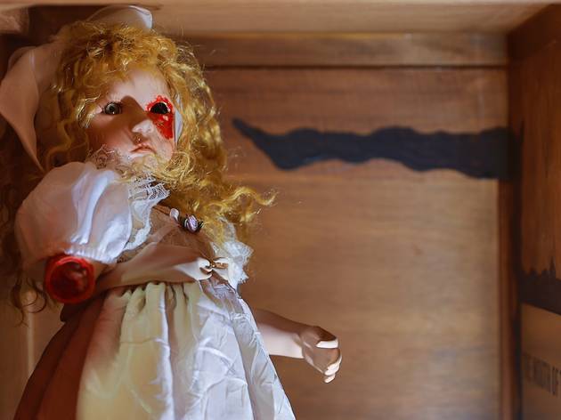 Escape or Die A porcelain doll wearing a white dress and with long curly blonde hair in a wooden box. The doll is missing an arm and an eye and red paint has been used to look like blood on the missing body parts