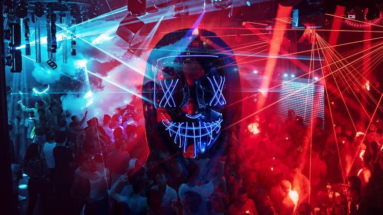A person wearing an LED mask and hood is silhouetted against a dance floor with lasers and smoke.