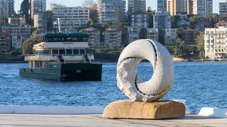 A granite ring-like sculpture by the harbour with a ferry in the background