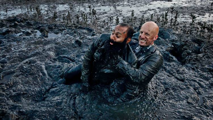 Two men wearing suits wrestle in a muddy pool