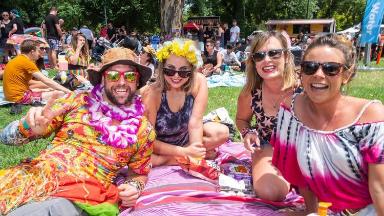 Colourfully dressed people in sunglasses, floral necklaces and head pieces smile at the camera as they sit on the grass in the sun.