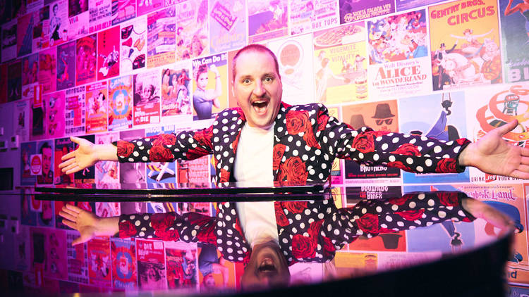 A person in a spotty and rose patterned blazer sits behind a piano with their arms wide open.