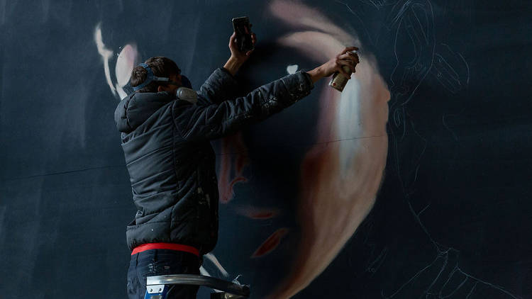 A man wearing a dark hooded jacket and a respiration mask created a mural of a woman's face using aerosol paint