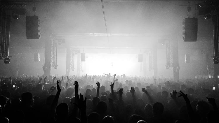 Large crowd of people inside a vast warehouse facing towards a lighted stage