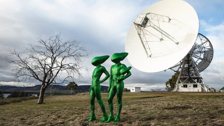 Two people wearing full body sparkly green suits with conical heads. They look like aliens and are standing in front of a huge satellite dish on a hillside