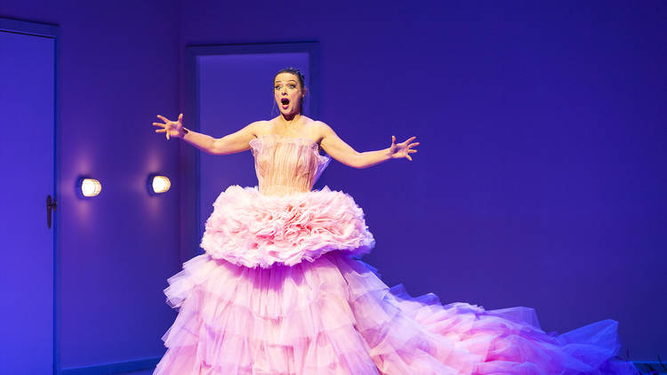 A woman in a floor-length tulle pink dress appears to sing loudly in a small, blue-lit room