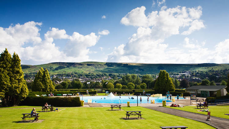 Green lawns with picnic benches dotted around them, in front of the bright blue pool at Ilkley Lido, with moors rising up behind it and a bright cloudy sky