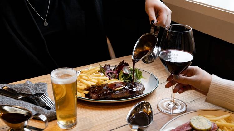 View of a table with food. A hand pours gravy on a steak with chips.