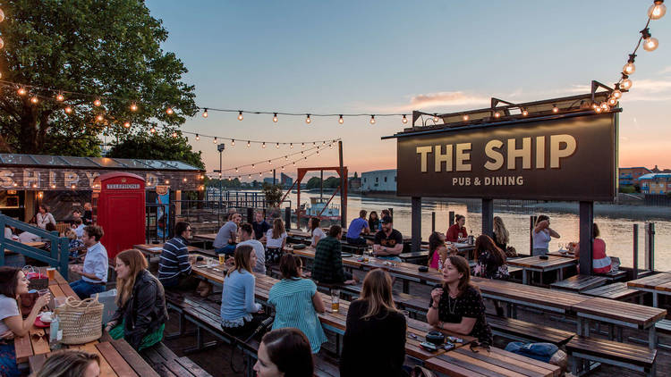 Groups of people sat at long wooden benches in a beer garden, with trees on the left, rows of lightbulbs overhea, a large sign saying 'The Ship Pub and Dining' and a pink sunset over the river Thames in the background