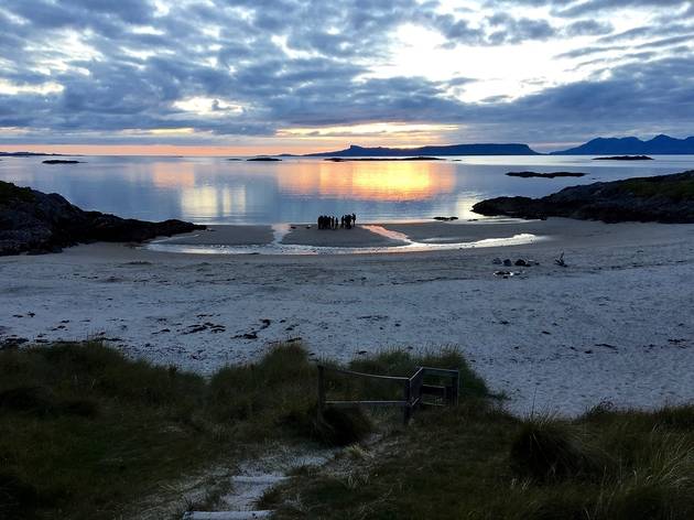 Camusdarach Beach in Scotland at Sunset A cloudy sky at sunset over a blue-tinged beach at low tide, with mountains in the far distance and scrub at the edge of the beach in the foreground